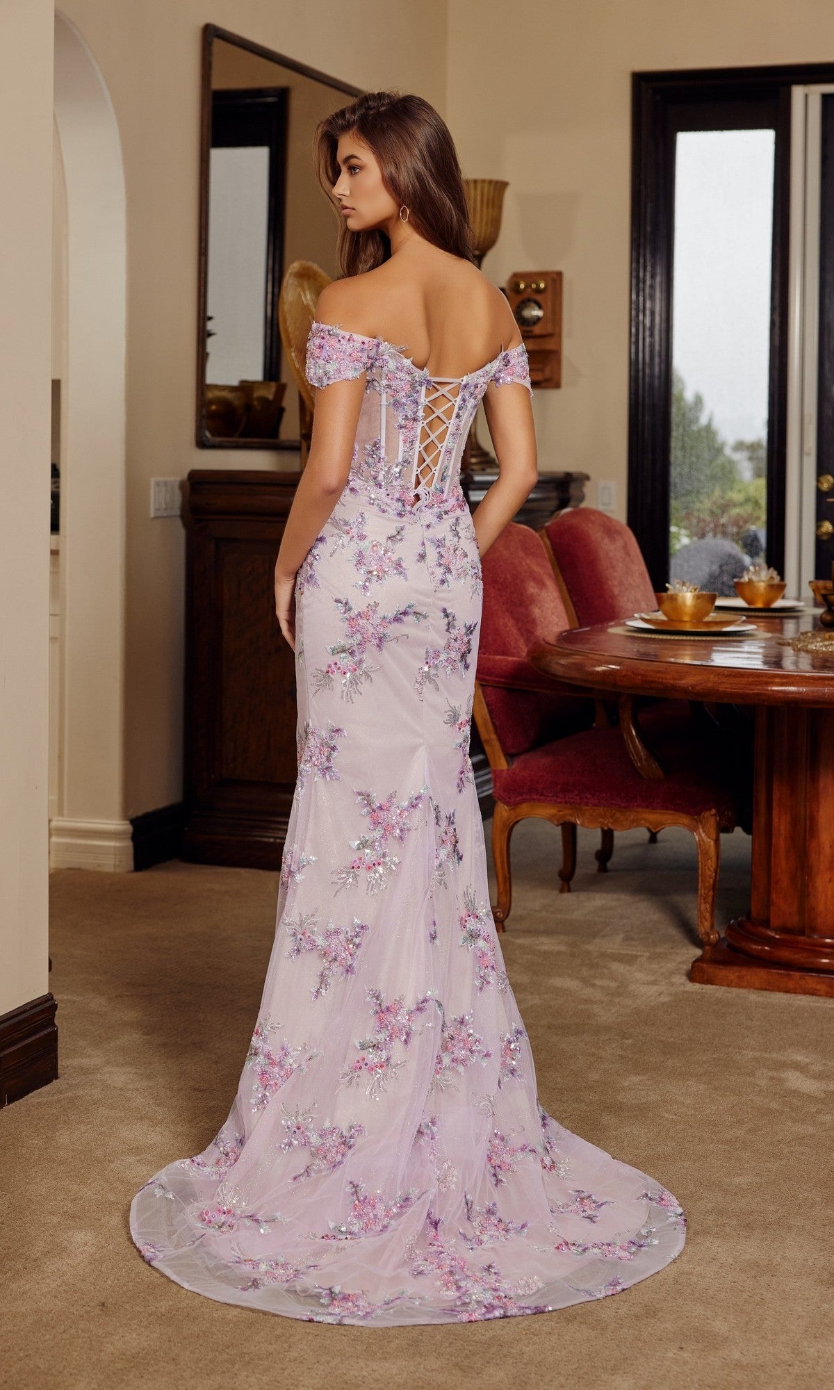 A woman indoors faces away in the Nox Anabel C1829 long prom dress—an off-the-shoulder, light purple gown with sequin floral embroidery and a corset bodice.